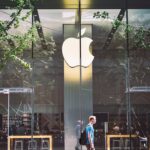 A casual scene of a man walking past the iconic glass facade of an Apple Store in Beijing, China.