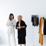 Two women discussing fashion business strategy with a laptop and clothes rack.