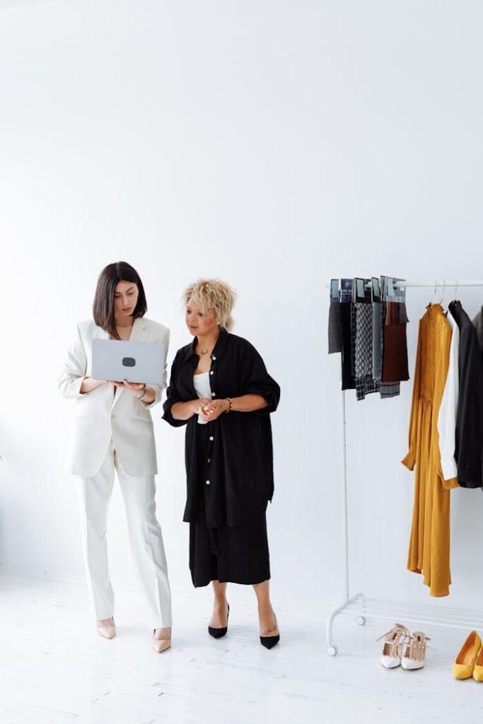 Two women discussing fashion business strategy with a laptop and clothes rack.